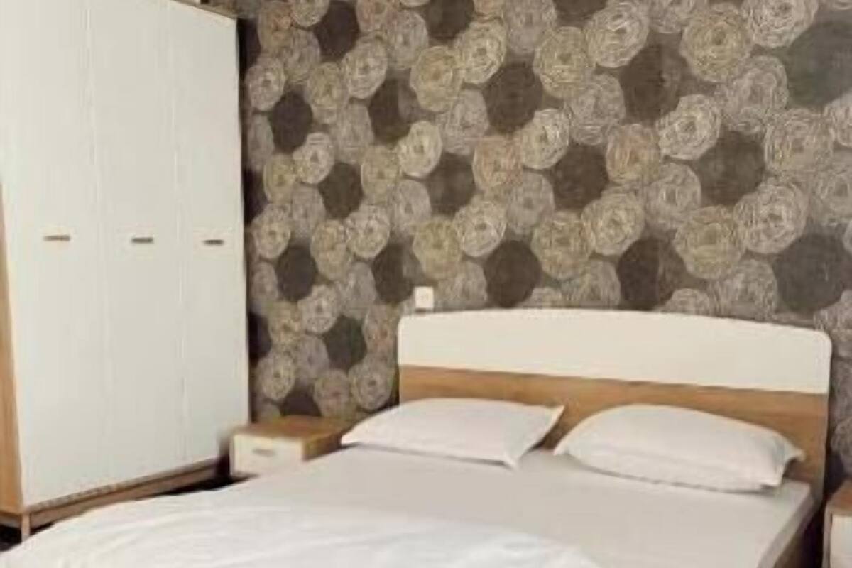 Una cama bien hecha con estructura de madera y ropa de cama blanca, un armario blanco y papel tapiz estampado.