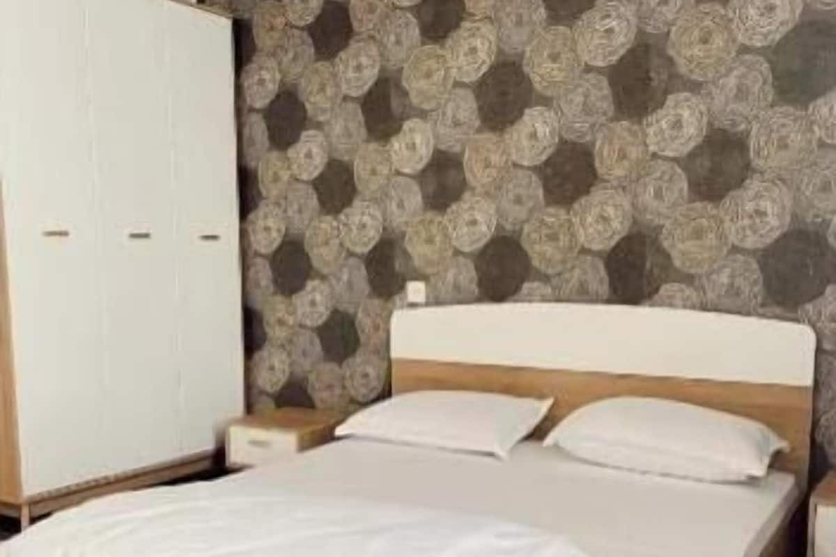 A neatly made bed with a wooden frame and white bedding, a white wardrobe, and patterned wallpaper.