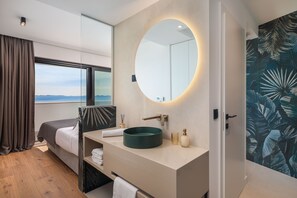 Ein modernes Hotelzimmer mit einem großen Bett, einem runden Spiegel, einem Waschbecken und Meerblick.