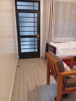 Ein Zimmer mit einem Bett, einem Holzsessel, einem Tisch, einem Fenster mit Vorhängen und einer Tür mit Gittermuster.