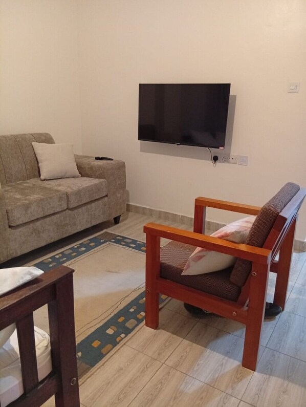 Uma sala de estar com sofá, uma cadeira de madeira, televisão fixada na parede e um tapete estampado.