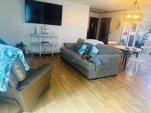 A living room with a grey sofa, a TV mounted on the wall, a chandelier, and a dining area with a table and chairs.