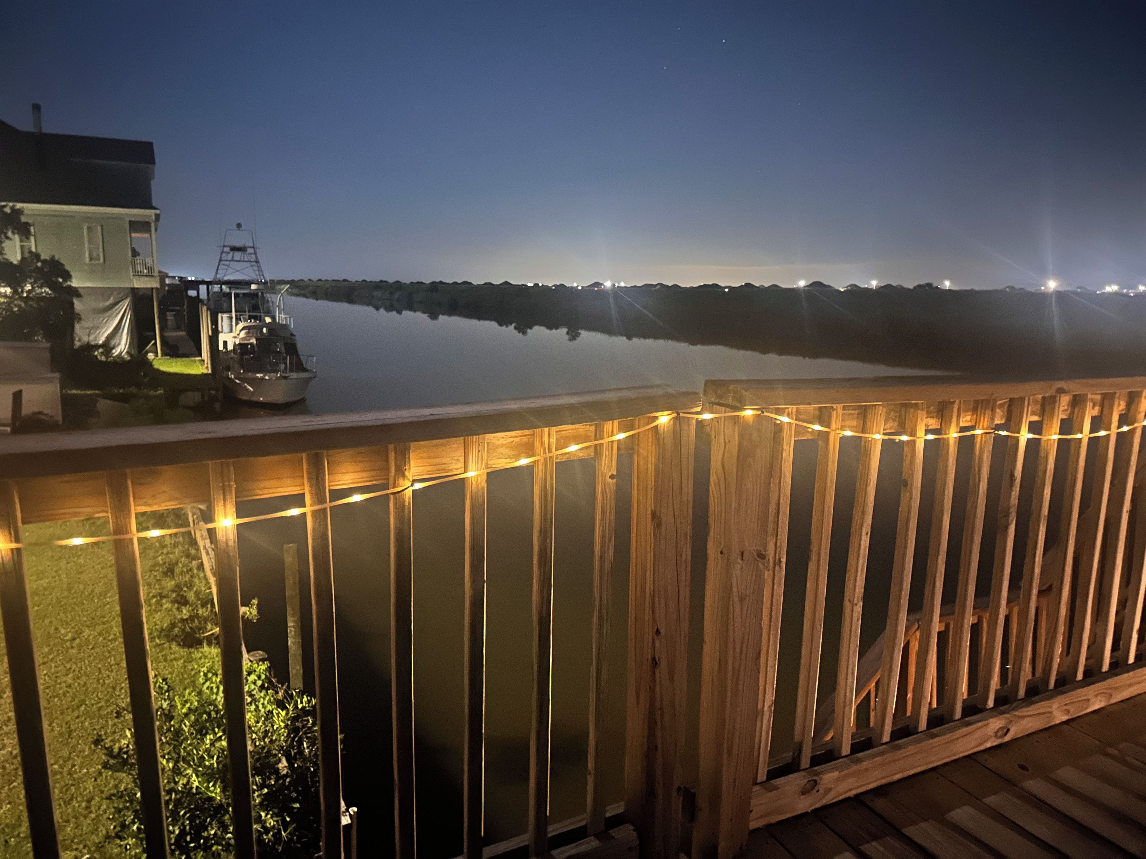 Une terrasse en bois, illuminée de guirlandes lumineuses, donnant sur une étendue d’eau calme la nuit.