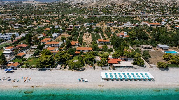 Luchtbeeld van een kustgebied met woonhuizen, een strand en een groot wit gebouw met blauwe parasols.