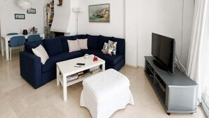 Een woonkamer met een donkerblauwe bank, een witte salontafel, een tv-meubel en een schilderij aan de muur.
