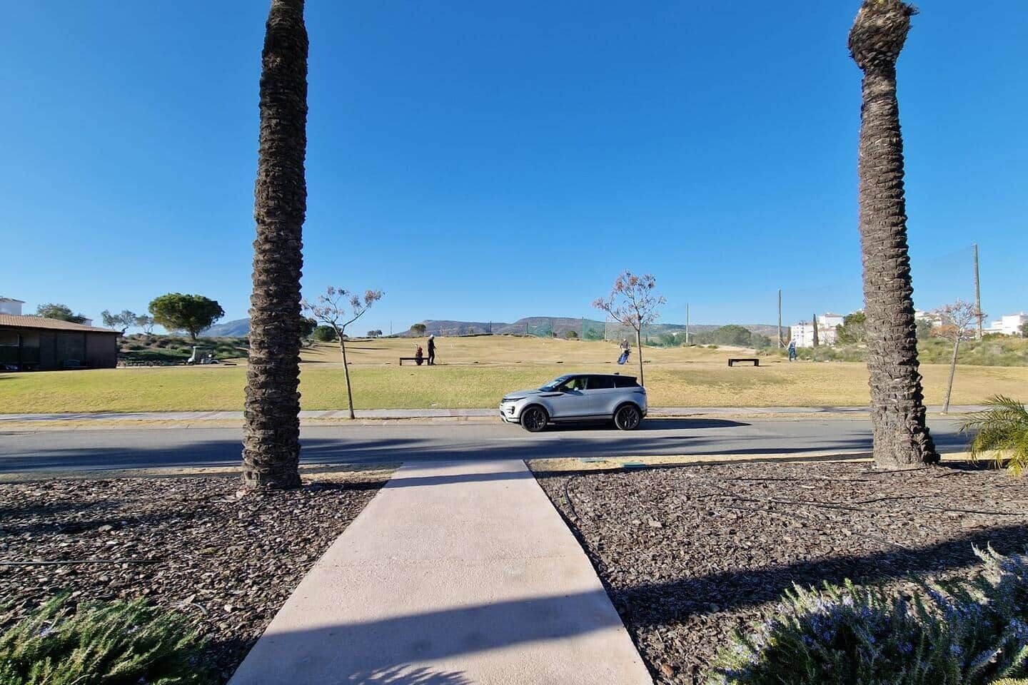 Un chemin pavé mène à un parking où se trouve un véhicule, des palmiers et un ciel dégagé.