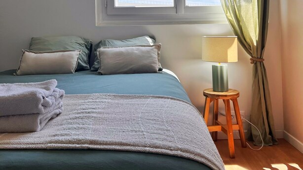 Een slaapkamer met een bed, kussens, een nachtkastje met een lamp, een raam met gordijnen en uitzicht op de lucht.