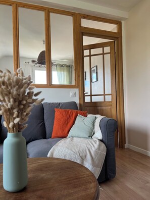 A living room with a sofa, a round wooden table, and a vase with dried flowers. - 10° Ouest - T3 renovated 2025 - Park view - Quiet street (Lorient)