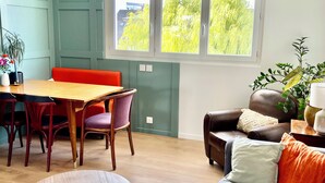 Een eetgedeelte met een houten tafel en stoelen, een bank met kussens, en een raam met uitzicht op bomen.