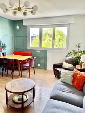 A dining area with a wooden table and chairs, a sofa with cushions, and a window with a view of trees. - 10° Ouest - T3 renovated 2025 - Park view - Quiet street (Lorient)