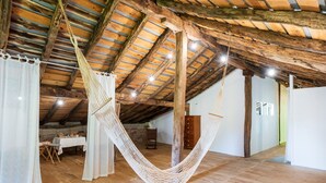 A hammock suspended from a wooden ceiling in a rustic room with a table and chairs, wooden floors, and a stone wall.