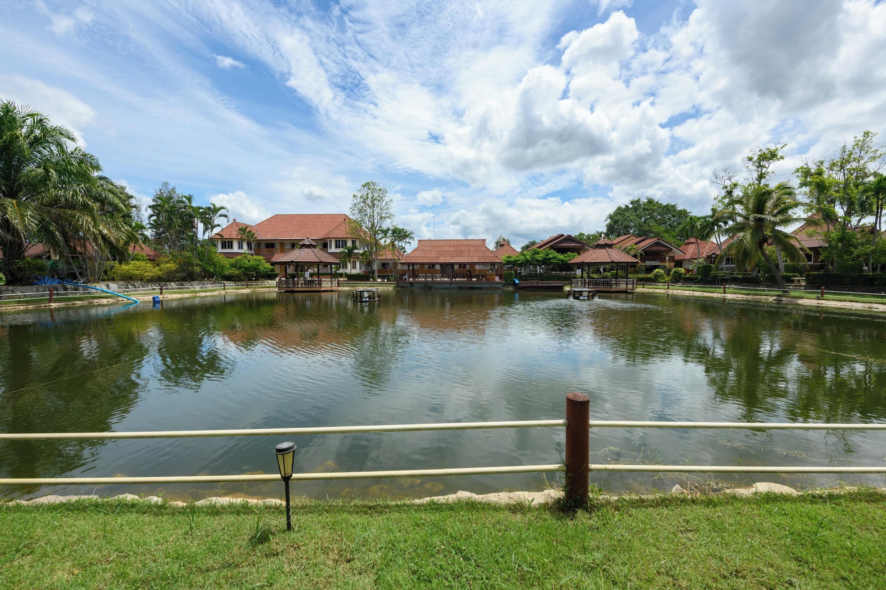 A serene lake with a wooden dock, surrounded by lush greenery and traditional-style buildings.