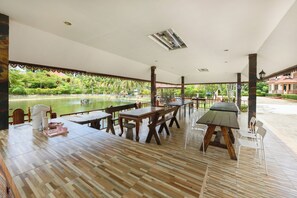 Un área de comedor al aire libre con mesas y sillas, vista a un cuerpo de agua, y un edificio de fondo.