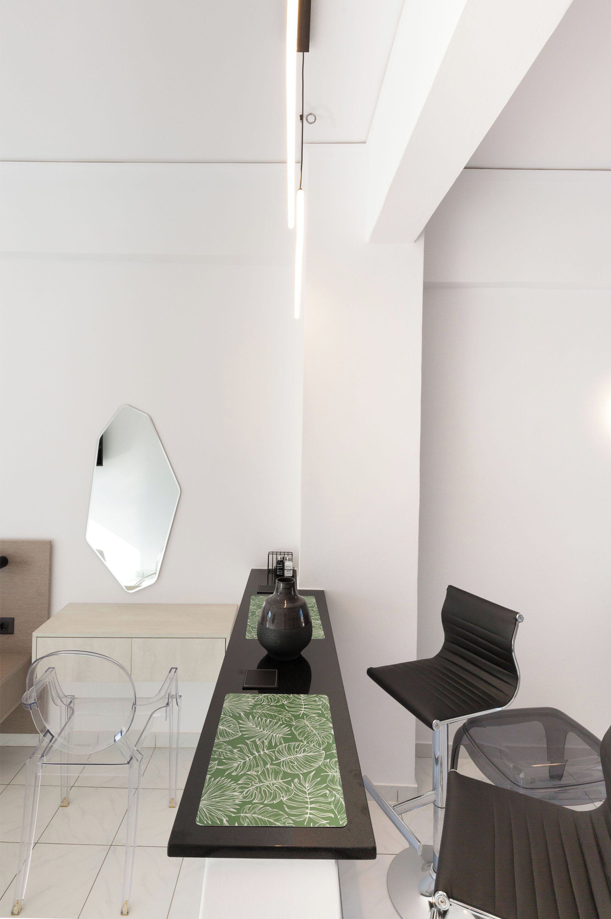 A modern, minimalist room with a long table, clear chairs, a black chair, a decorative mirror, and a green patterned table runner.
