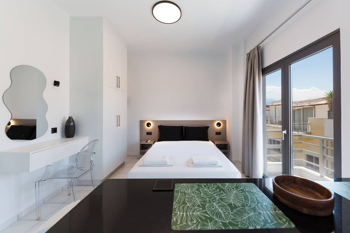 Una habitació d'hotel moderna amb un llit gran, taules de nit, un mirall, un escriptori amb una cadira i vista a la ciutat a través de grans finestres.