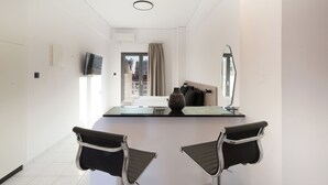 A modern hotel room with a minimalist design, featuring a large mirror, a flat-screen TV, a bedside table with a vase, and two black chairs.