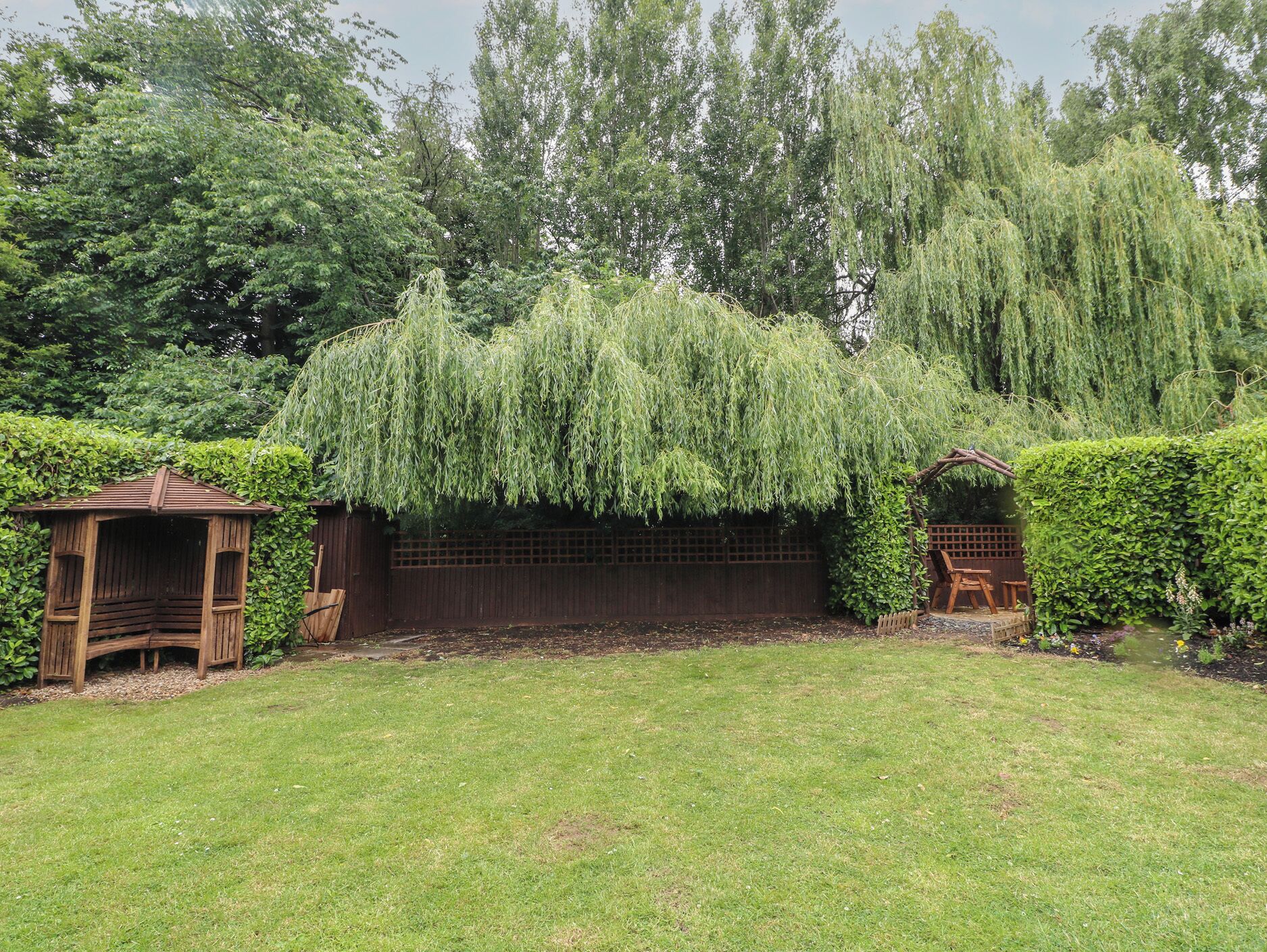 Un jardin agrémenté d’un kiosque en bois, d’un saule pleureur et d’un mur en briques.