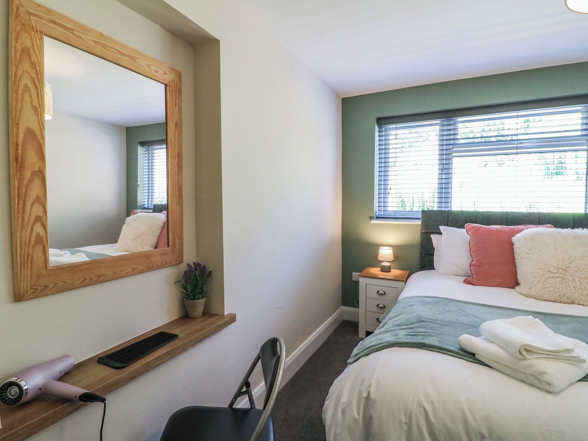 Een slaapkamer met een bed, nachtkastje, lamp, spiegel, stoel en een plank met een haardroger en toetsenbord.