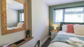 Een slaapkamer met een bed, nachtkastje, lamp, spiegel, stoel en een plank met een haardroger en toetsenbord.