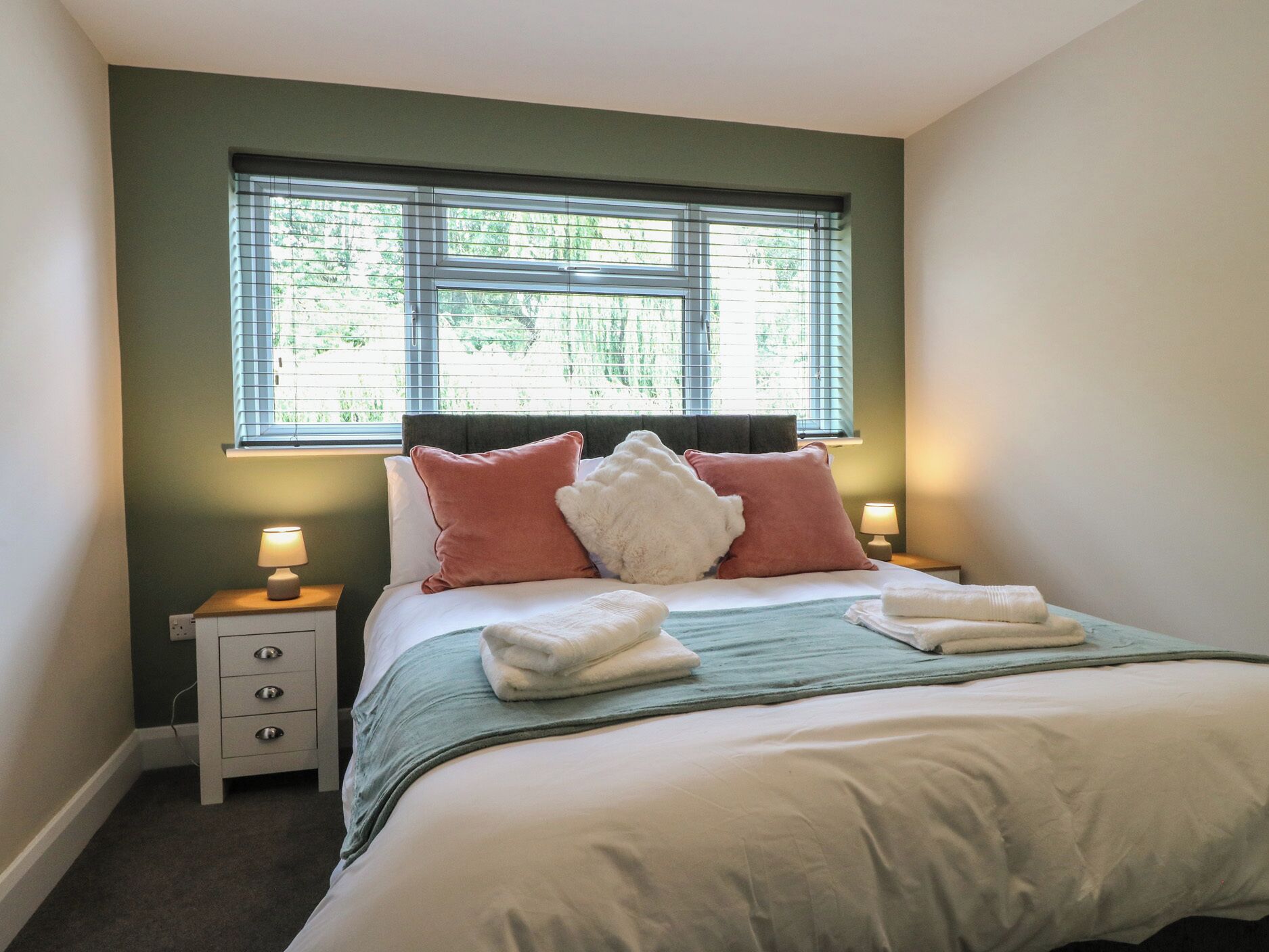 A neatly made bed with pillows and a blanket, a bedside table with a lamp, and a window with blinds.