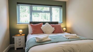 A neatly made bed with pillows and a blanket, a bedside table with a lamp, and a window with blinds.