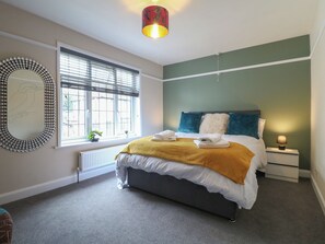 Ein Schlafzimmer mit einem Bett, einem Fenster mit Jalousien, einem Nachttisch mit Lampe und einem an der Wand befestigten Spiegel.