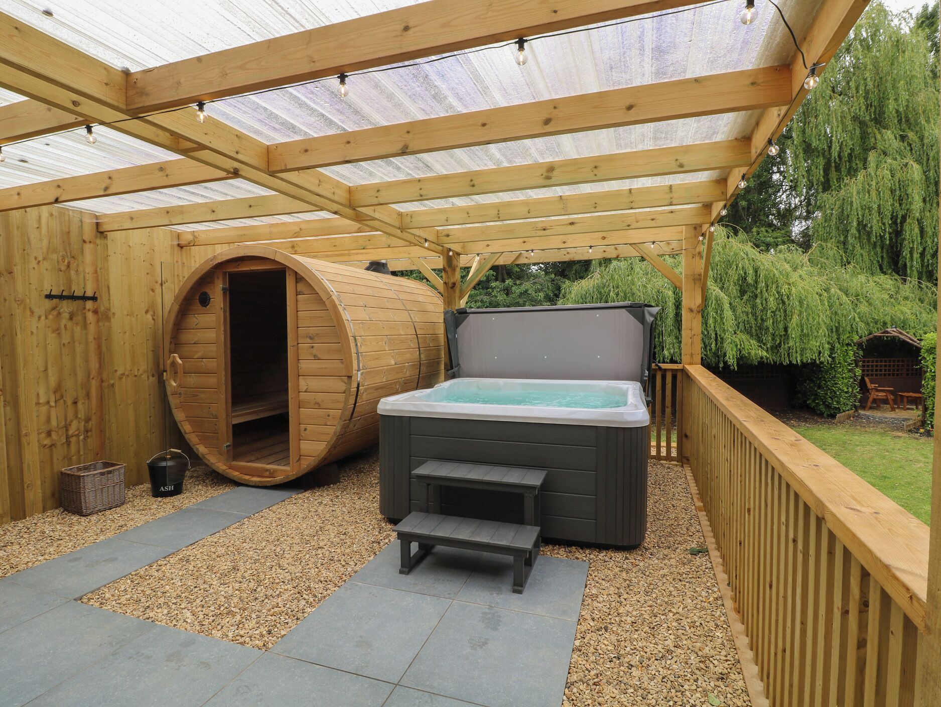 A wooden spa area with a hot tub, a wooden barrel, and a pergola with string lights.