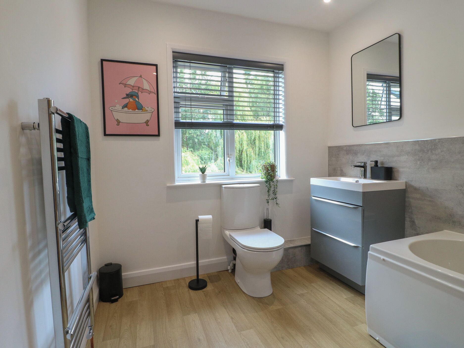 Un baño moderno con inodoro, lavabo y bañera, un radiador, una ventana con persianas y un cuadro en la pared.