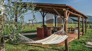 Eine Hängematte unter einer Holz-Pergola, ein Backstein-Pflanzgefäß und ein Tisch mit Stühlen in einer grünen Außenanlage.