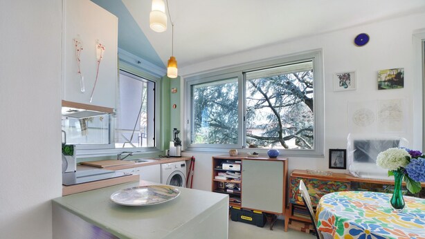Une cuisine équipée d’un lave-linge, une table à manger recouverte d’une nappe colorée et une fenêtre donnant sur des arbres.