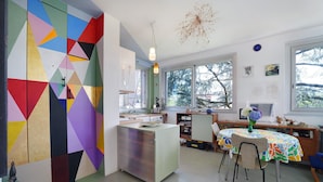 Une cuisine avec un mur aux motifs géométriques et aux couleurs vives, une table à manger avec des chaises, et une vue dégagée grâce à de grandes fenêtres.