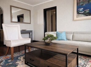 Un salón con un sofá blanco, un sillón blanco, una mesa de centro de madera oscura, una planta en maceta y un cuadro de barcos.
