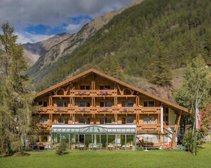 Una cabaña de madera con varios balcones, un invernadero de cristal y un paisaje montañoso de fondo.