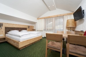 A room with a wooden bed, a desk, a chair, a TV mounted on the wall, and a window with sheer curtains.