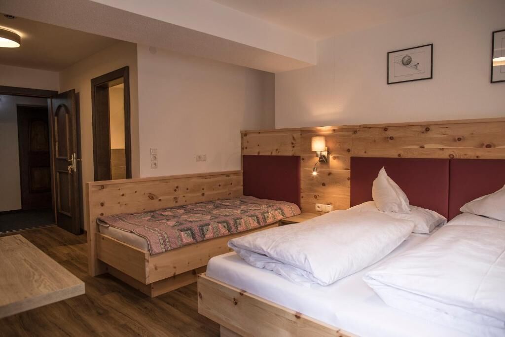A room with a wooden bed, a smaller bed with a patterned blanket, a wooden headboard, a wall-mounted lamp, and a door leading to another room.