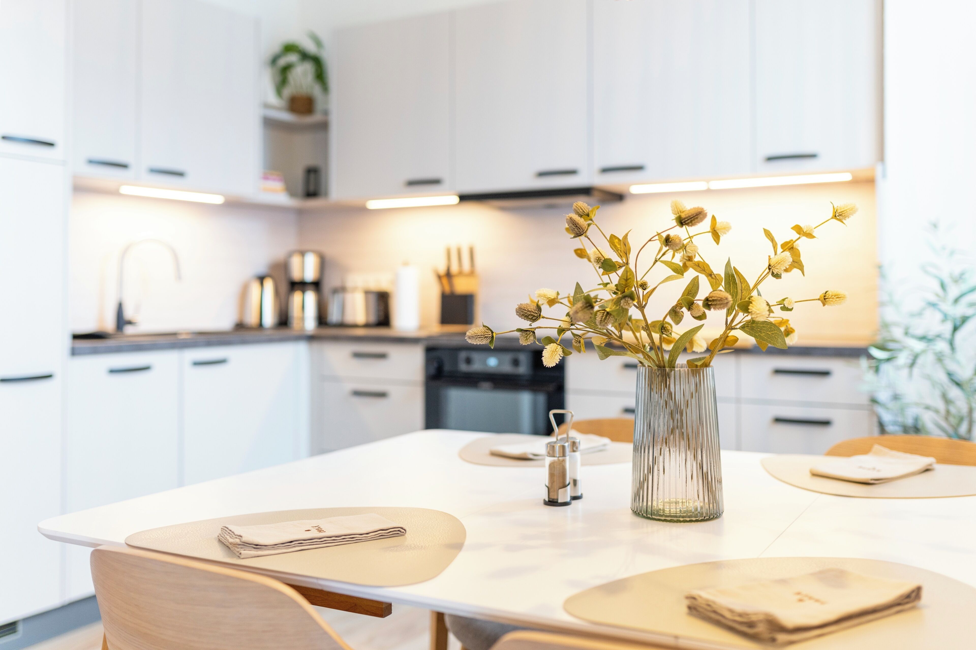A modern kitchen with white cabinets, a dining table with placemats, and a vase with flowers.
