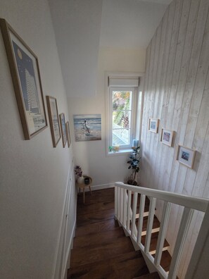 A staircase with wooden steps and white railings, leading to a room with a window and framed artwork on the walls.