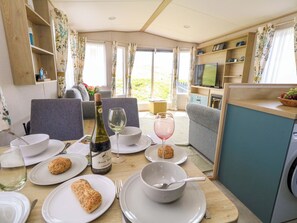 A dining area with a table set for a meal, a bottle of wine, and a television in the background. - Caravan A7 (Bridport)