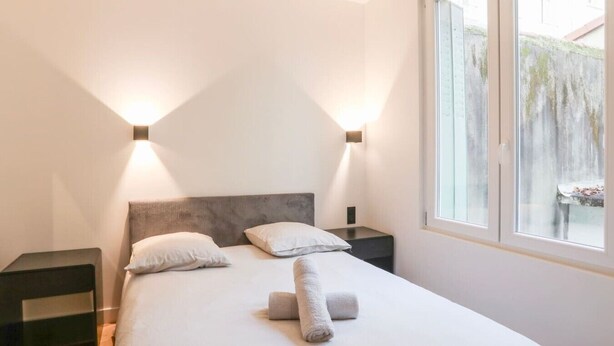 Un dormitorio con una cama, dos mesitas de noche, un ventanal y dos lámparas de pared.