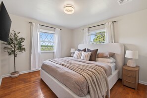 Une chambre à coucher avec un grand lit, deux fenêtres avec des rideaux, une table de chevet avec une lampe et une plante en pot.