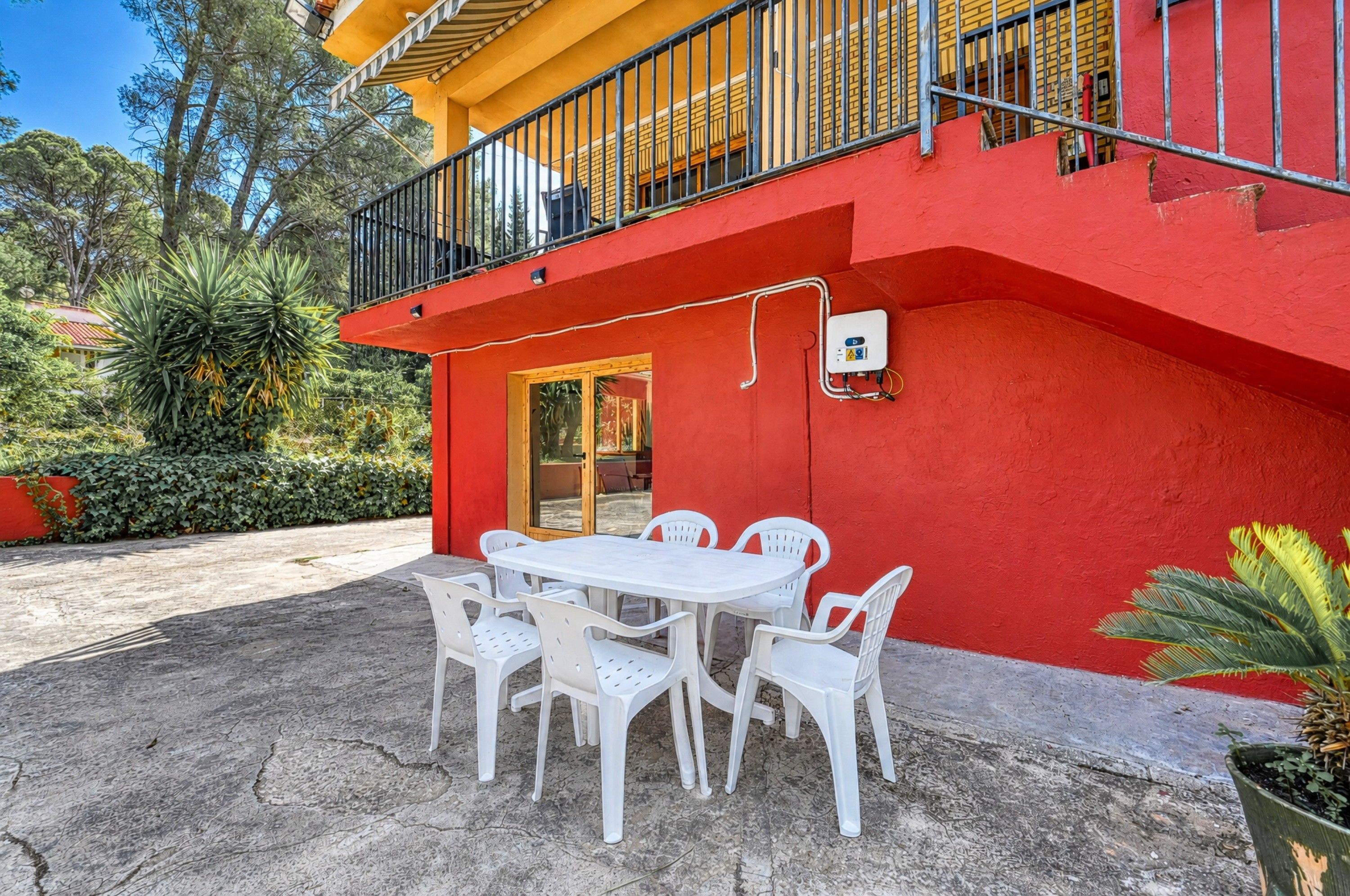 Un edificio rojo con un balcón, una mesa blanca con sillas y una planta en maceta.
