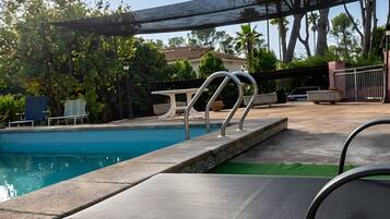 Una piscina con una escalera de metal, una zona de estar cubierta y una vista de un edificio al fondo.