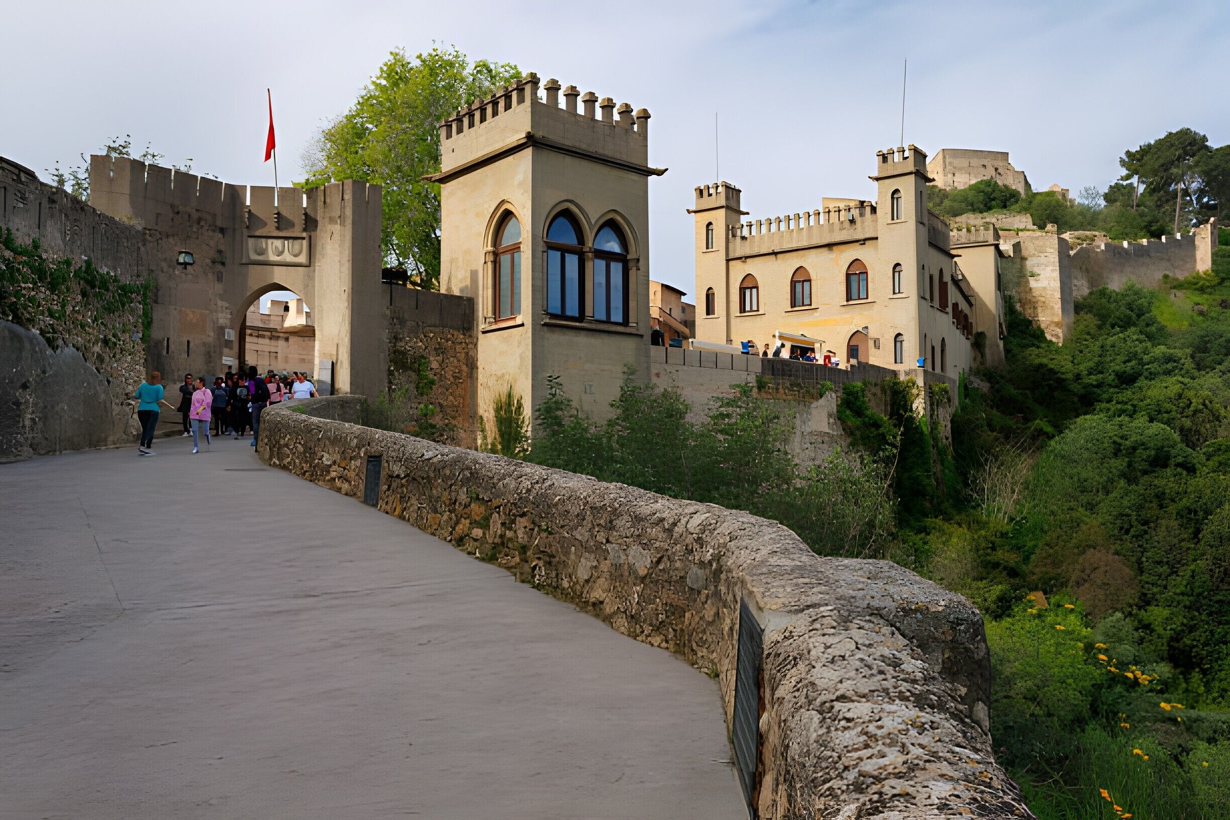 Un puente de piedra histórico que conduce a un castillo con ventanas de arco y una bandera.