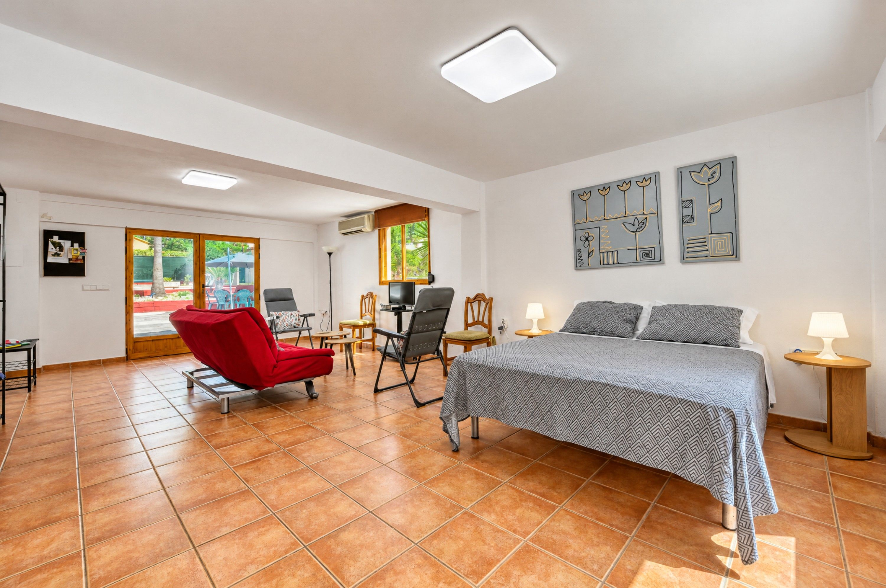 Una habitación amplia con una cama, una silla roja, un escritorio con computadora y un ventanal con vista a follaje.