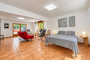 Une pièce spacieuse comprenant un lit, un fauteuil rouge, un bureau avec un ordinateur et une fenêtre donnant sur un paysage verdoyant.