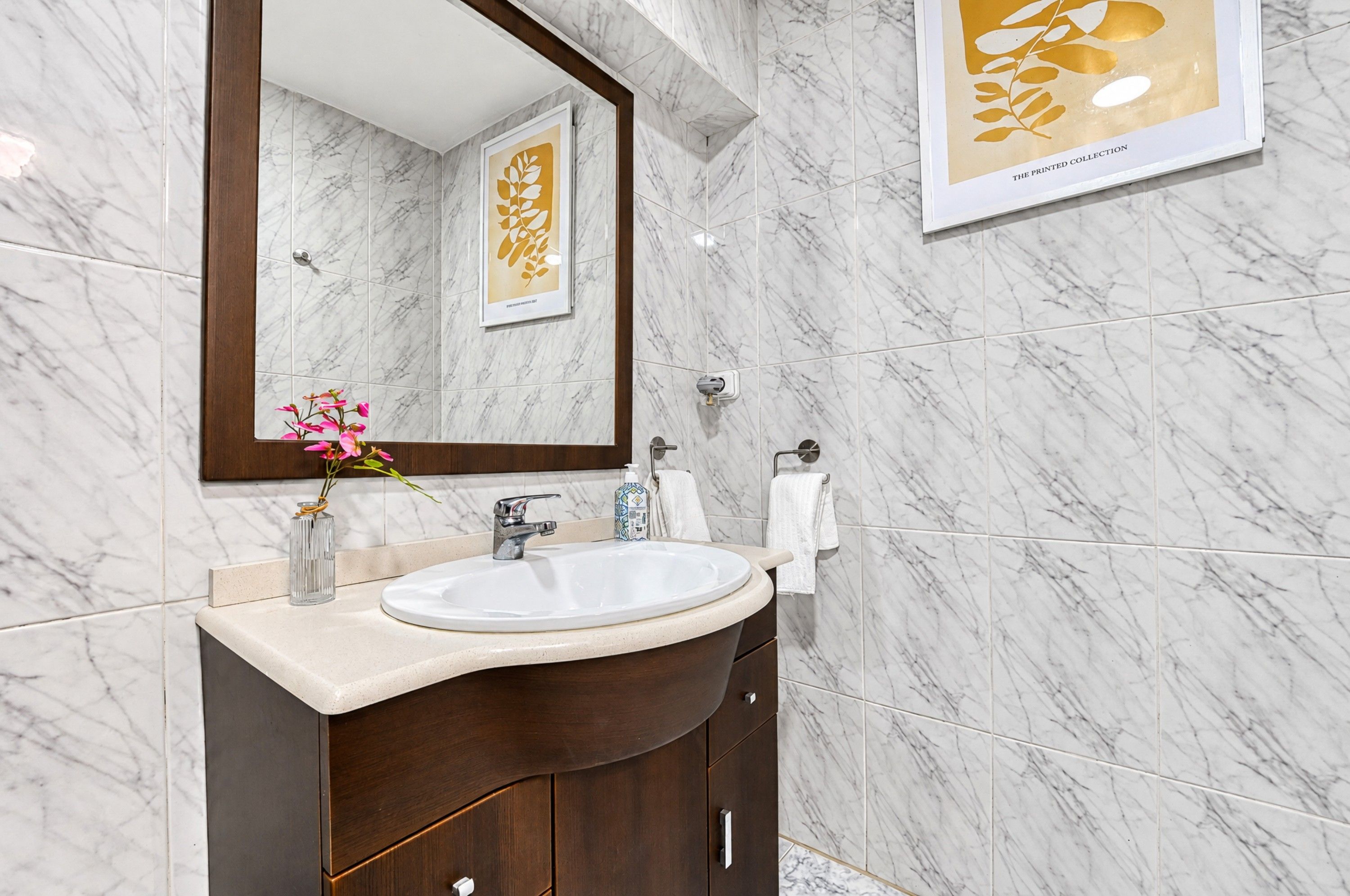 A bathroom with a marble wall, a large mirror, a sink, a vase with flowers, and a framed artwork.