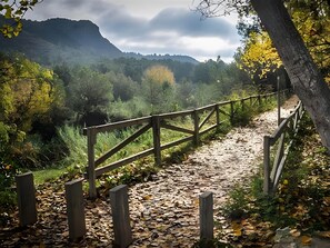 A wooden pathway with a fence, surrounded by trees and a mountain in the background. - Holiday Home 'Cova Negra Nature' with Mountain Views, Shared Pool, and Air Conditioning (El Genovés)