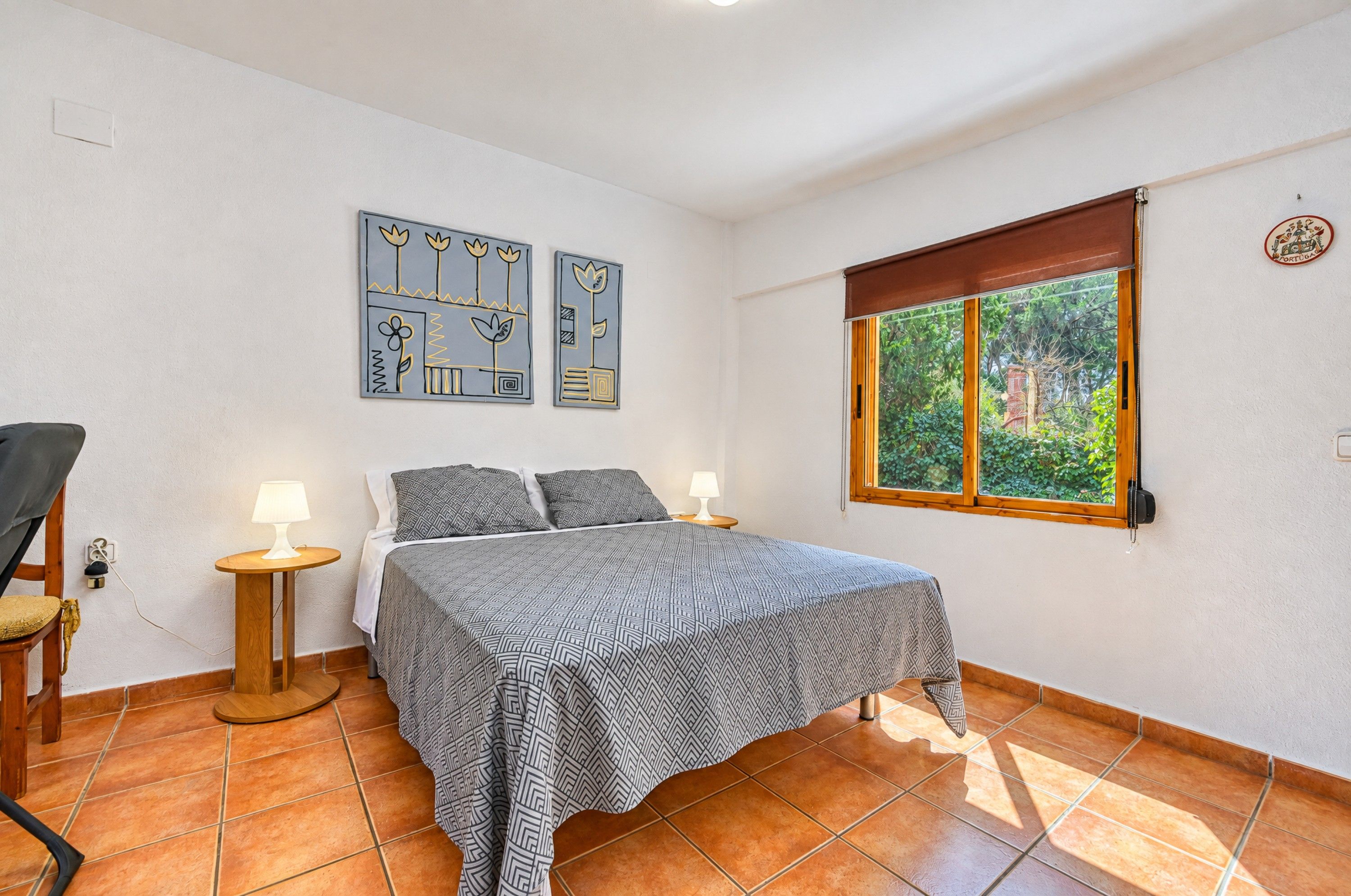 Un dormitorio con cama, dos mesitas de noche, una silla, una ventana con vistas a zonas verdes y dos obras de arte en las paredes.