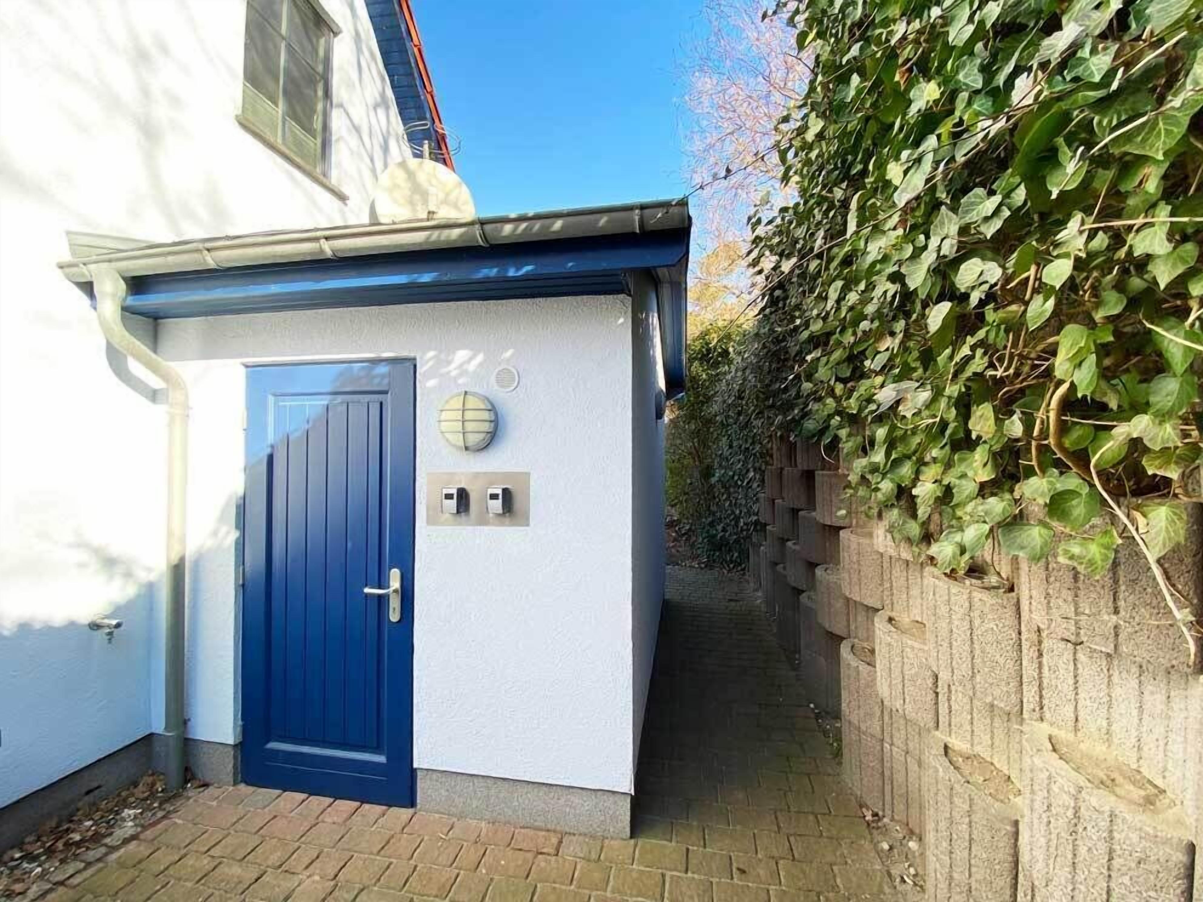 A small, white building with a blue door, a window, and a wall-mounted light fixture.