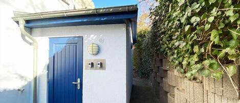A small, white building with a blue door, a window, and a wall-mounted light fixture.